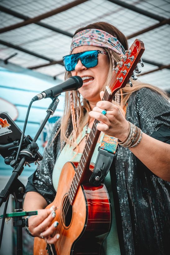 Fox Maple singing into a microphone while playing an acoustic guitar, wearing sunglasses, a patterned headband and a sequined jacket