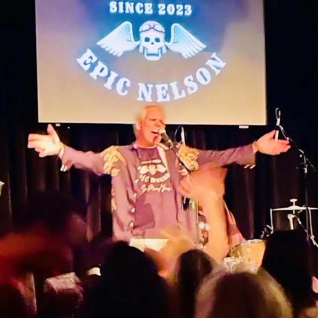 EPIC NELSON founder and lead vocalist Eric Nelson singing with arms outstretched on stage in front of the band's winged skull backdrop