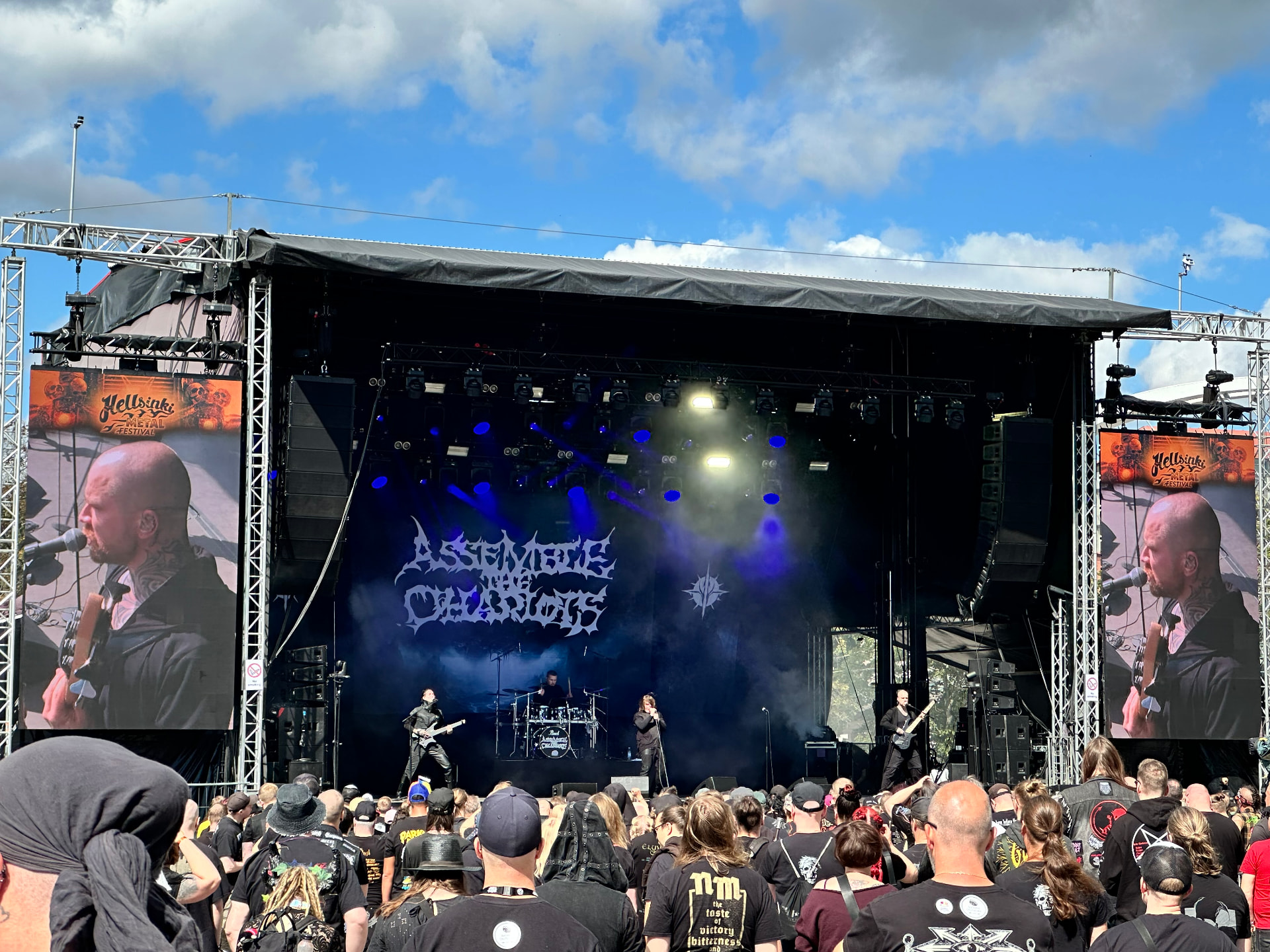 Assemble the Chariots — opening our Day Two. Assemble the Chariots performing on the outdoor stage at Hellsinki Metal Festival 2026 under a blue sky with their logo on the backdrop
